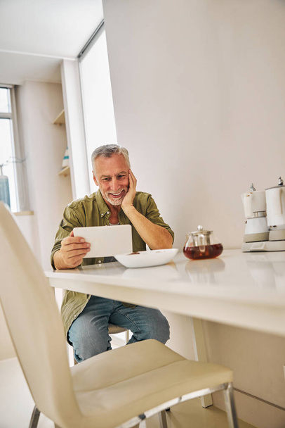 Smiley aged gentleman looking at tablet screen during teatime - Fotó, kép