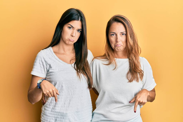 Famille hispanique de mère et fille portant un t-shirt blanc décontracté pointant vers le bas regardant triste et contrarié, indiquant la direction avec les doigts, malheureux et déprimé.  - Photo, image