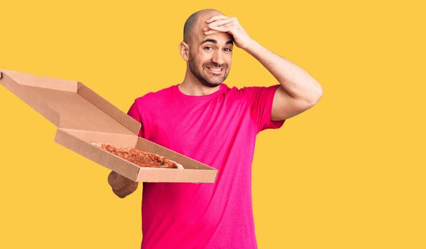 Νεαρός όμορφος άνδρας κρατώντας παράδοση πίτσα κουτί από χαρτόνι τόνισε και απογοητευμένος με το χέρι στο κεφάλι, έκπληκτος και θυμωμένος πρόσωπο  - Φωτογραφία, εικόνα
