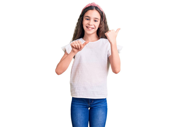 Mignon hispanique enfant fille portant un t-shirt blanc décontracté pointant vers le dos derrière avec la main et les pouces vers le haut, souriant confiant  - Photo, image