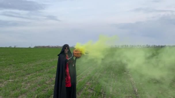 Young woman stands among field in Halloween costume of death with painted skeleton on her body and holds jack-o-lantern in her hands with yellow smoke from it. Halloween concept. Sugar skull makeup. - Footage, Video