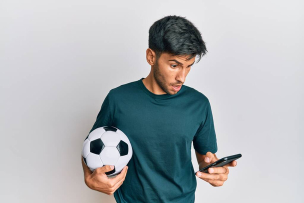 Młody przystojny mężczyzna trzyma piłkę nożną patrząc na smartphone bet app w twarz szoku, patrząc sceptyczny i sarkastyczny, zaskoczony otwartymi ustami  - Zdjęcie, obraz
