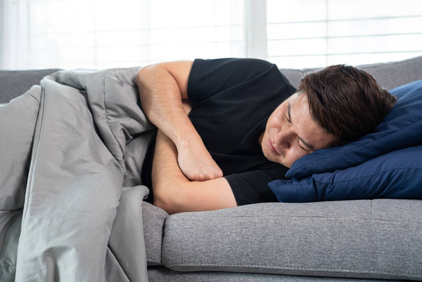 Zieke en onwel Aziatische mannen houden zijn hoofd, hebben een sterke hoofdpijn en slapen op de bank thuis verpakt in grijze deken. - Foto, afbeelding