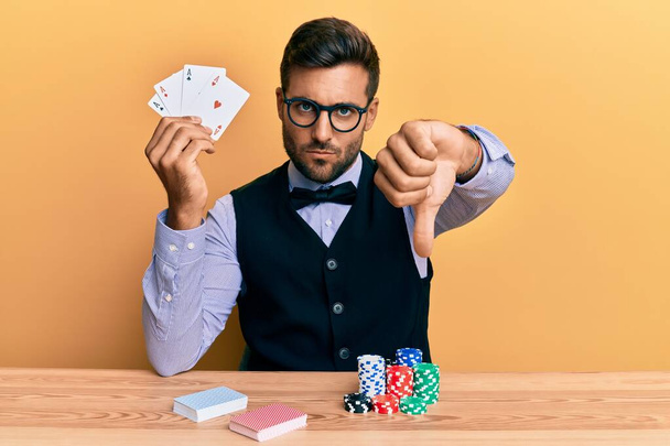 Bonito croupier hispânico homem sentado na mesa com fichas de poker e cartas com cara zangada, sinal negativo mostrando antipatia com os polegares para baixo, conceito de rejeição  - Foto, Imagem
