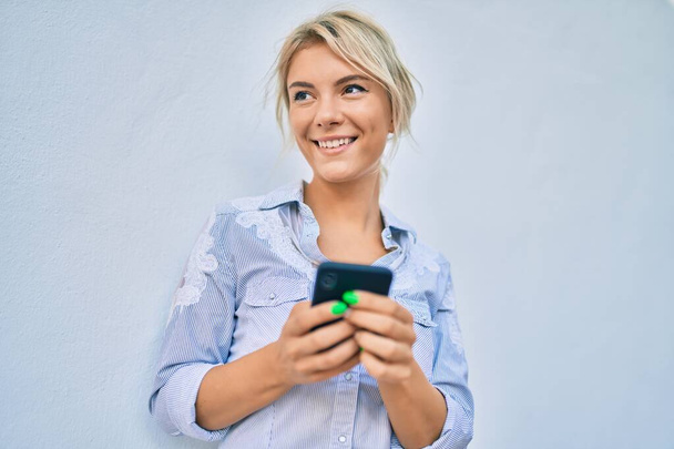Молода блондинка посміхається щаслива, використовуючи смартфон у місті
. - Фото, зображення