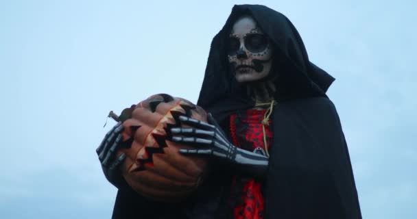 Jovem mulher fica no traje de Halloween da morte com esqueleto pintado em seu corpo e jack-o-lanterna contra o fundo do céu. Conceito de Halloween. Maquiagem do crânio açúcar. - Filmagem, Vídeo