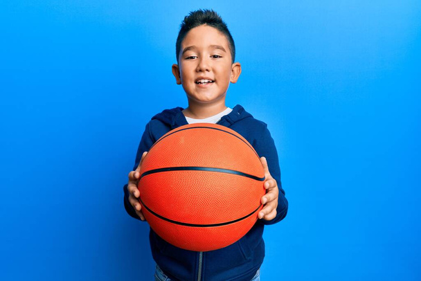 Malý kluk, hispánec držící basketbal, se usmívá se šťastným a chladným úsměvem na tváři. zobrazování zubů.  - Fotografie, Obrázek