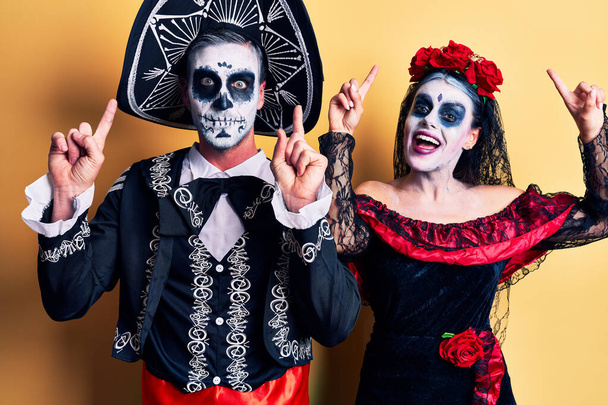 Молодая пара, одетая в мексиканский день мертвого костюма над желтой улыбкой удивлена и удивлена и указывает пальцами и поднятыми руками.  - Фото, изображение