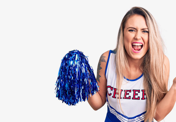 Young beautiful blonde woman wearing cheerleader uniform holding pompom screaming proud, celebrating victory and success very excited with raised arms  - Photo, Image