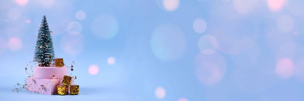 Trendy Christmas and New Year banner with Christmas tree, present boxes, decorations and pink bokeh lights on pastel blue background with copyspace. Selective focus, place for text - Photo, Image