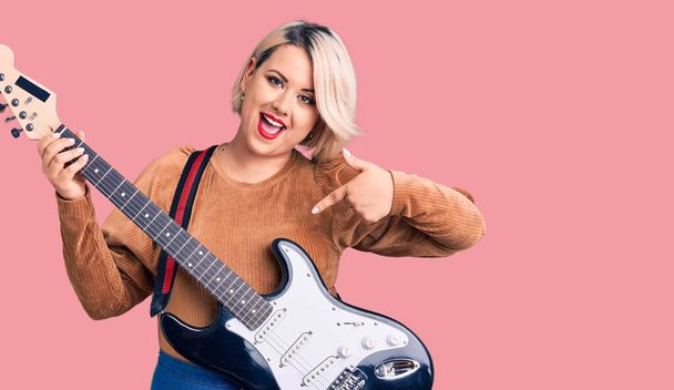Young blonde plus size woman playing electric guitar smiling happy pointing with hand and finger  - Photo, Image