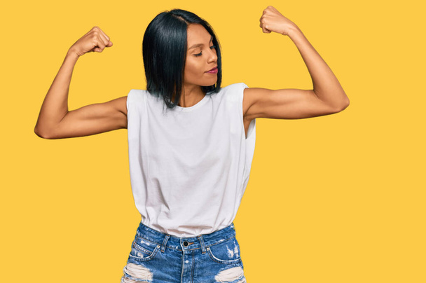 Молодая африканская американка, одетая в повседневную одежду, показывает мышцы рук, улыбающиеся с гордостью. фитнес концепция.  - Фото, изображение