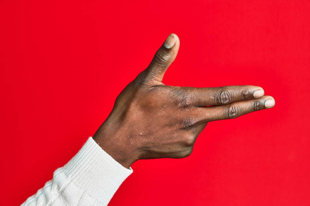 Bras et main de jeune homme noir afro-américain sur fond rouge isolé gesticulant arme à feu avec les doigts, visant symbole de tir  - Photo, image