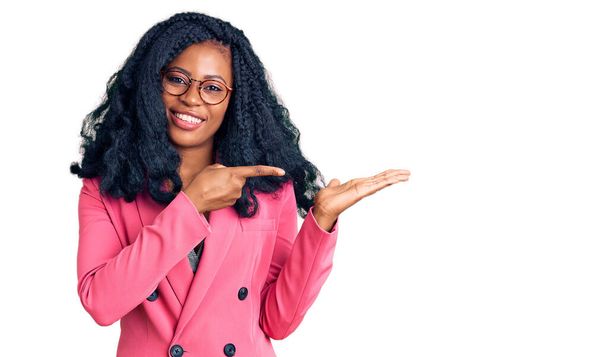 Прекрасна афро-американська жінка, одягнена в бізнес-піджак і окуляри вражені і посміхаючись до камери, представляючи з рукою і вказуючи пальцем.  - Фото, зображення