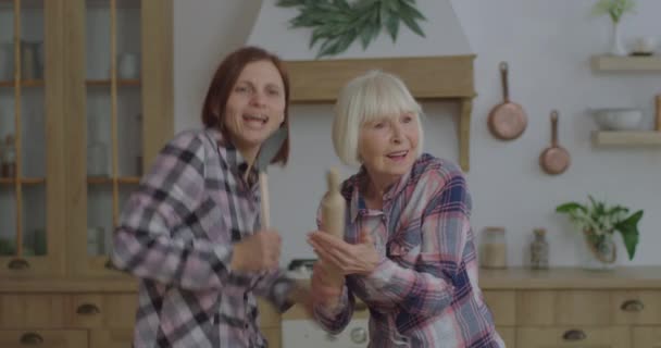 Happy family of young adult woman and senior mother dancing and singing into kitchenware like a microphone at kitchen. Women having fun with rolling pin and scoop staying at home. - Filmagem, Vídeo