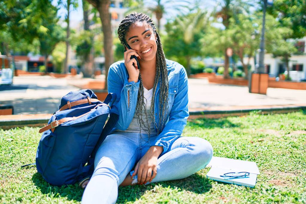 Jovem estudante afro-americana sorrindo feliz falando no smartphone sentado na grama no campus da universidade - Foto, Imagem