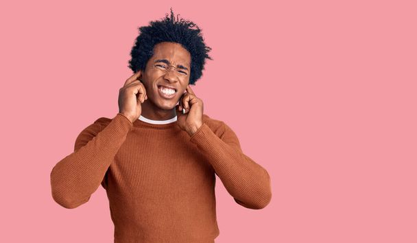 Schöner afrikanisch-amerikanischer Mann mit Afro-Haaren, der lässig gekleidet ist und die Ohren mit Fingern bedeckt, die sich über den Lärm lauter Musik ärgern. Gehörloses Konzept.  - Foto, Bild
