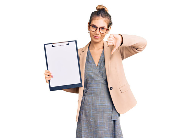 Beautiful caucasian woman with blonde hair holding clipboard with blank space with angry face, negative sign showing dislike with thumbs down, rejection concept  - Fotoğraf, Görsel