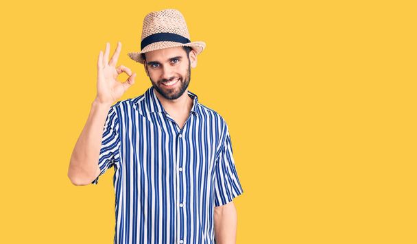 Homem bonito jovem com barba usando chapéu de verão e camisa listrada sorrindo positivo fazendo sinal ok com a mão e os dedos. expressão bem sucedida.  - Foto, Imagem