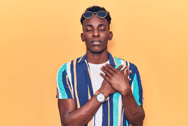 Young african american man wearing summer clothes and sunglasses smiling with hands on chest with closed eyes and grateful gesture on face. health concept.  - Fotó, kép