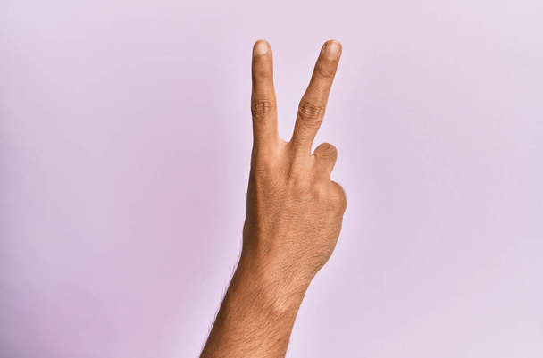 Brazo y mano del joven caucásico sobre fondo rosa aislado contando el número 2 mostrando dos dedos, señalando victoria y símbolo ganador  - Foto, Imagen