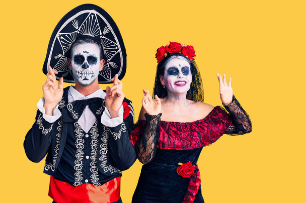Jong stel draagt Mexicaanse dag van het dode kostuum over achtergrond gebaar vinger gekruist glimlachend met hoop en ogen dicht. geluk en bijgelovig concept.  - Foto, afbeelding