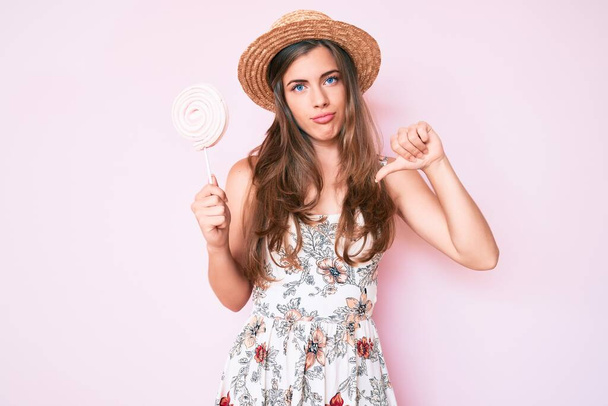 Beautiful young caucasian woman wearing summer hat and lollipop with angry face, negative sign showing dislike with thumbs down, rejection concept  - Foto, afbeelding