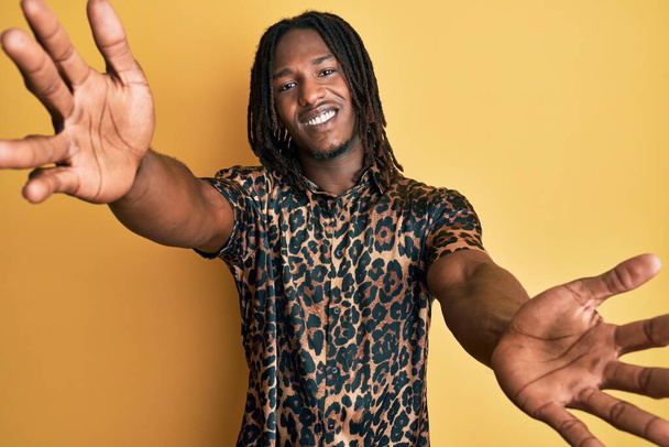 Afrikanischer Mann mit Zöpfen im Leoparden-Animal-Print-Hemd, der lächelnd mit offenen Armen in die Kamera blickt, um sich zu umarmen. fröhlicher Ausdruck, der das Glück umarmt.  - Foto, Bild