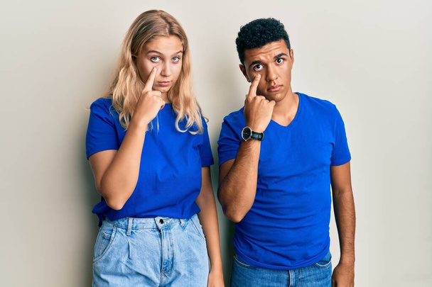 Νεαρό διαφυλετικό ζευγάρι φοράει καθημερινά ρούχα που δείχνουν το μάτι βλέποντας σας χειρονομία, ύποπτη έκφραση  - Φωτογραφία, εικόνα