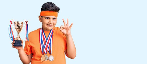 Niño pequeño sosteniendo trofeo campeón y usando medallas haciendo signo ok con los dedos, sonriendo gesto amistoso excelente símbolo  - Foto, imagen