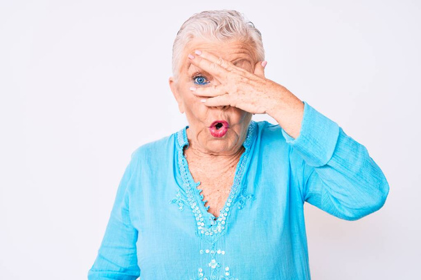 Starší krásná žena s modrýma očima a šedivými vlasy v letních šatech vykukující v šoku zakrývající obličej a oči rukou, dívající se skrz prsty  - Fotografie, Obrázek