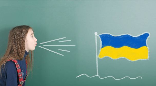 Niña pre-adolescente soplando en el tablero de la escuela dibujado en la bandera de Ucrania pizarra. Foto de alta resolución. Profundidad total del campo.  - Foto, Imagen