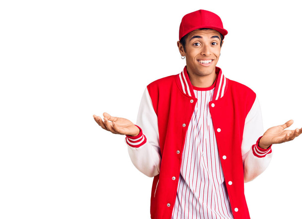 Молодой афроамериканец, одетый в бейсбольную форму, ничего не знает и путает выражения лица с поднятыми руками и руками. Концепция сомнений.  - Фото, изображение