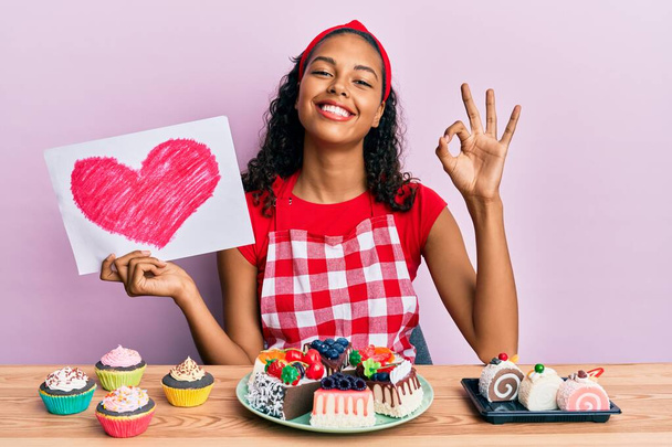 Young african american girl wearing baker uniform holding heart draw sitting on the table with sweets doing ok sign with fingers, smiling friendly gesturing excellent symbol  - 写真・画像