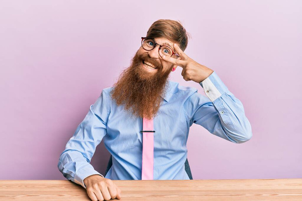 Молодой ирландский рыжий мужчина в деловой рубашке и галстуке сидит на столе и делает символ мира с пальцами на лице, улыбаясь весело показывая победу  - Фото, изображение