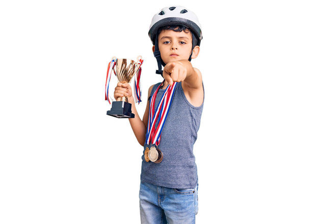 小さなかわいい男の子は自転車のヘルメットを身に着けていると受賞者のトロフィーを保持するメダルは、カメラとあなたに指で指して、自信を持ってジェスチャーを真剣に見て  - 写真・画像