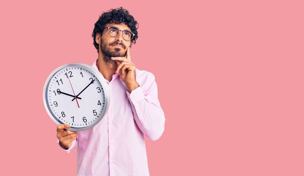 Handsome young man with curly hair and bear holding big clock serious face thinking about question with hand on chin, thoughtful about confusing idea  - Foto, Imagem