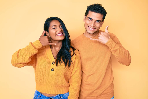 Beautiful latin young couple wearing casual clothes together smiling doing phone gesture with hand and fingers like talking on the telephone. communicating concepts.  - Photo, Image
