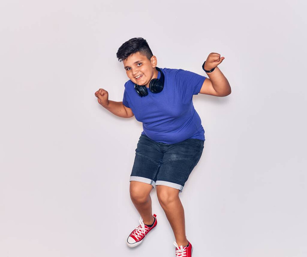 Adorable hispanic child boy smiling happy. Jumping with smile on face listening to music using headphones over isolated white background - Foto, Imagem