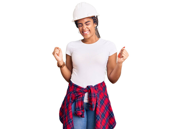 Jonge Afro-Amerikaanse vrouw met vlechten met een harde hoed en bouwer kleding erg blij en enthousiast doen winnaar gebaar met armen opgeheven, glimlachen en schreeuwen om succes. viering concept.  - Foto, afbeelding