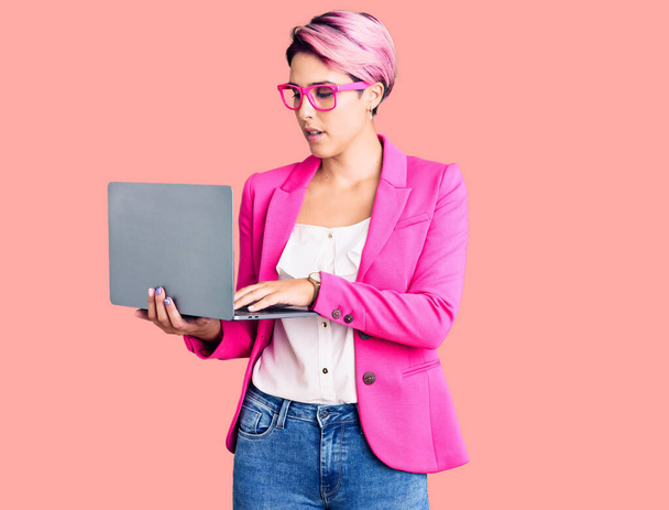 Giovane bella donna con i capelli rosa che indossa occhiali che tengono l'atteggiamento di pensiero del computer portatile e l'espressione sobria guardando sicuro di sé  - Foto, immagini