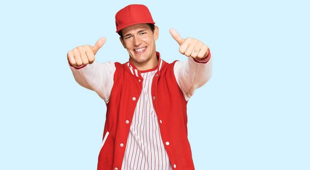 野球のユニフォームを着たハンサムな白人男性が手で積極的なジェスチャーをし、親指を上げて笑顔で成功に満足しています。勝者のジェスチャー.  - 写真・画像