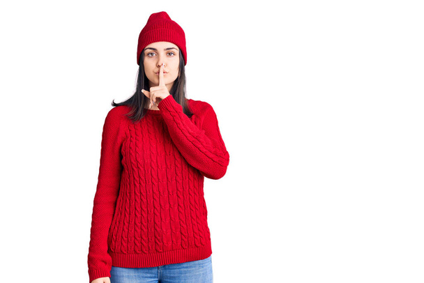Молодая красивая девушка в свитере и шерстяной шапке просит тишины с пальцем на губах. молчание и тайная концепция.  - Фото, изображение