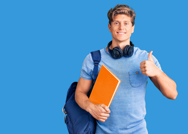 Jonge knappe man in studentenrugzak en koptelefoon met boek glimlachend en positief, duim omhoog doen uitstekend en goedkeuring teken  - Foto, afbeelding