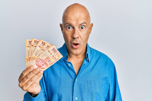 Middle age bald man holding mexican pesos scared and amazed with open mouth for surprise, disbelief face  - Photo, Image