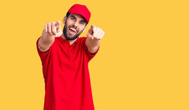 Young handsome man with beard wearing delivery uniform pointing to you and the camera with fingers, smiling positive and cheerful  - Zdjęcie, obraz