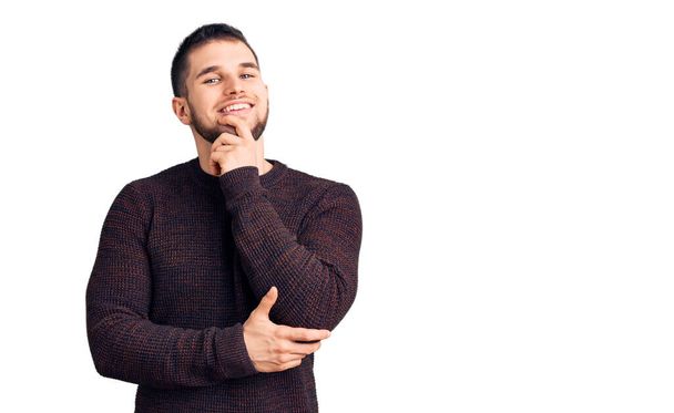 Young handsome man wearing casual sweater looking confident at the camera with smile with crossed arms and hand raised on chin. thinking positive.  - Foto, Imagem