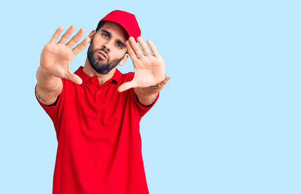 Young handsome man with beard wearing delivery uniform doing frame using hands palms and fingers, camera perspective  - Fotografie, Obrázek