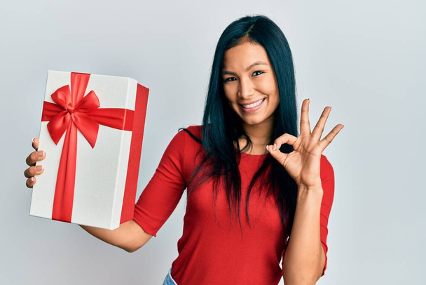 Beautiful hispanic woman holding gift doing ok sign with fingers, smiling friendly gesturing excellent symbol  - 写真・画像