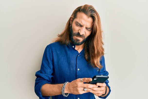 Attractive man with long hair and beard using smartphone skeptic and nervous, frowning upset because of problem. negative person.  - Foto, immagini